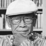 Older man wearing a white knit cap and glasses resting his chin on his hand in a library setting black and white photo